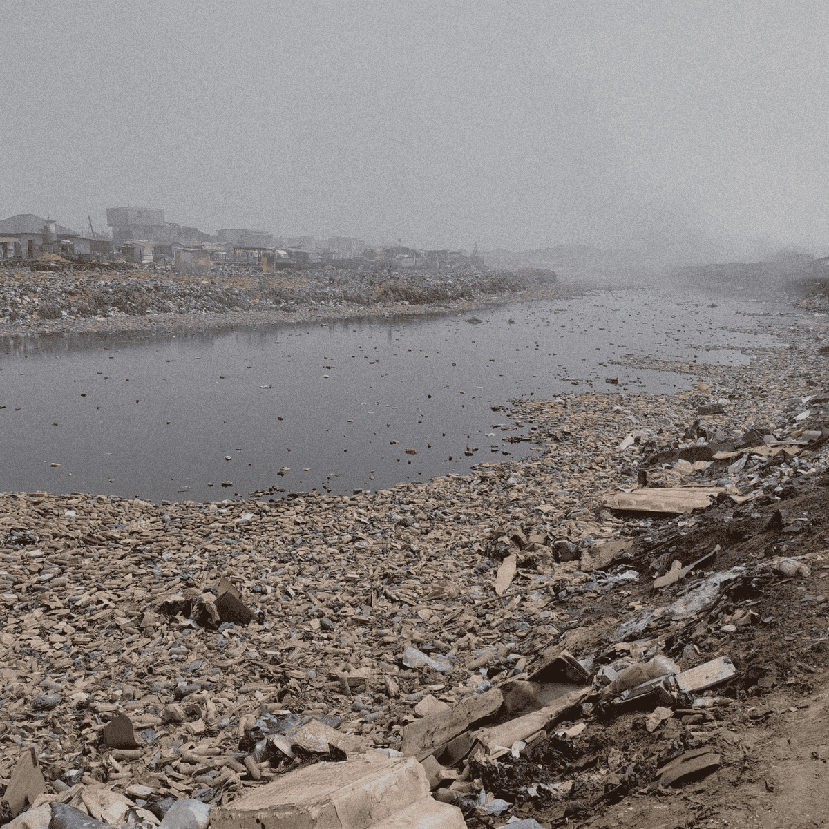 Polluted river with garbage and debris, surrounded by industrial buildings on a foggy day.